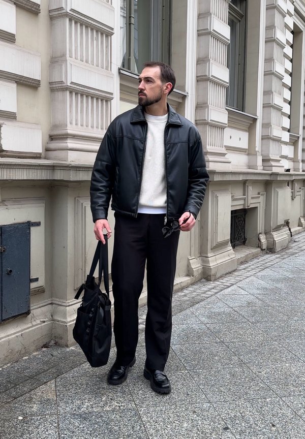 Homme en veste en cuir noire et pantalon noir tenant un sac noir et des lunettes de soleil, debout sur le trottoir de la ville près d'un bâtiment orné.