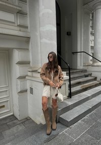 Brun cropped jakke, hvid blondekjole, beige broderede westernstøvler og en lille hvid håndtaske. Marmortrapper og tekstureret væg i baggrunden.