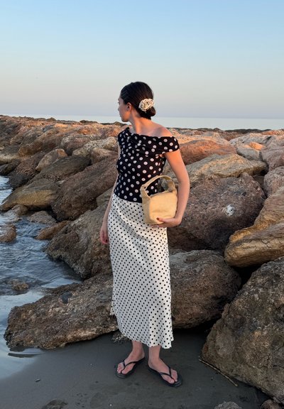 Top off-the-shoulder de lunares en blanco y negro combinado con una falda de lunares, sosteniendo un bolso de mimbre beige y usando chanclas negras.