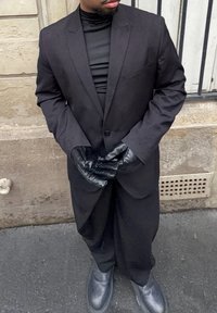 Costume noir avec un blazer cintré et un pantalon, porté sur un col roulé noir. Des gants et des chaussures noirs brillants complètent le look.