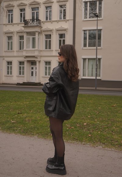 Chaqueta de cuero negra de gran tamaño con bolsillos laterales, combinada con medias negras transparentes y botas negras gruesas, con un edificio de fondo.