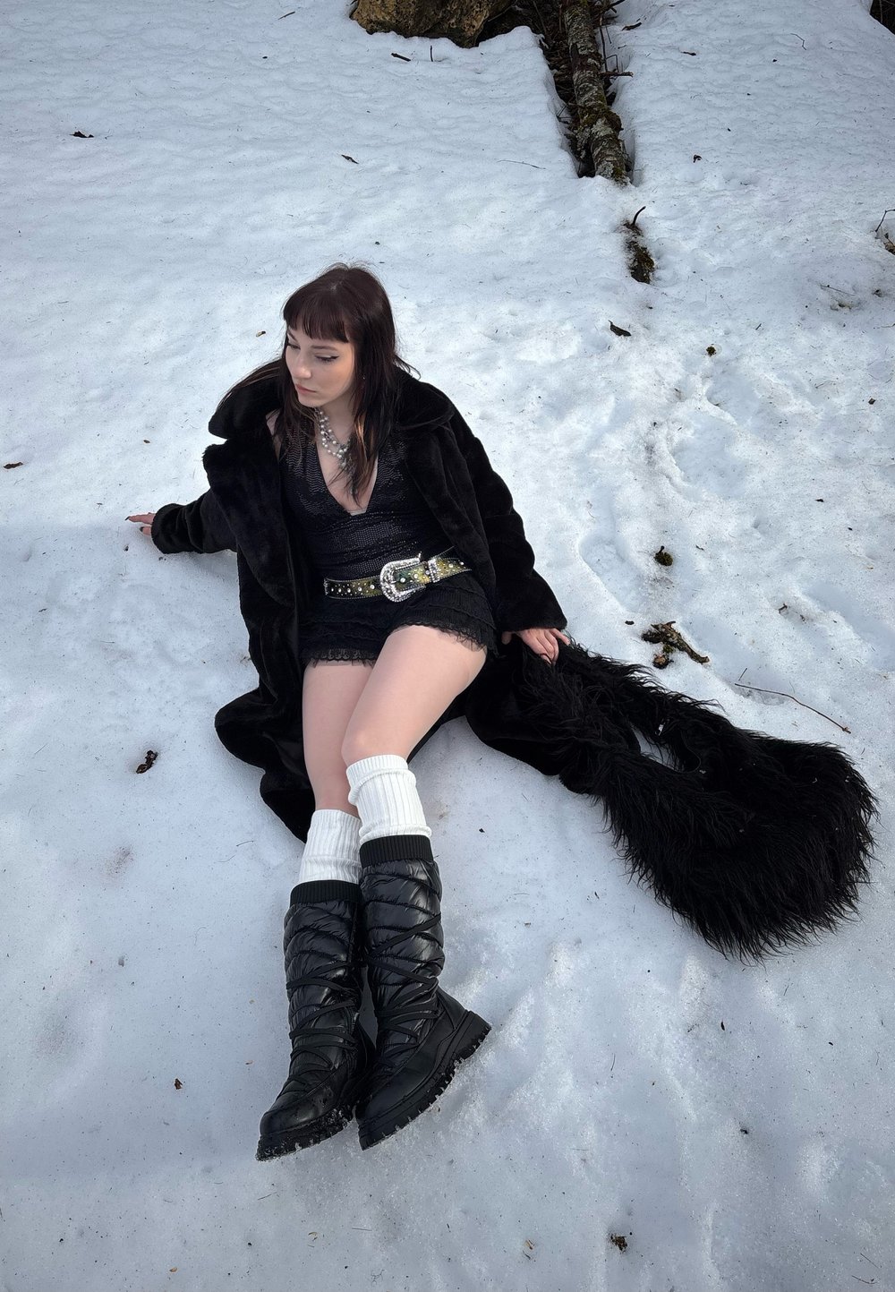 Cappotto in finta pelliccia nera, abito in pizzo nero e stivali neri alti fino al ginocchio con texture. Calze bianche fino al ginocchio, collana a strati e frange lunghe sul cappotto. Terreno innevato.