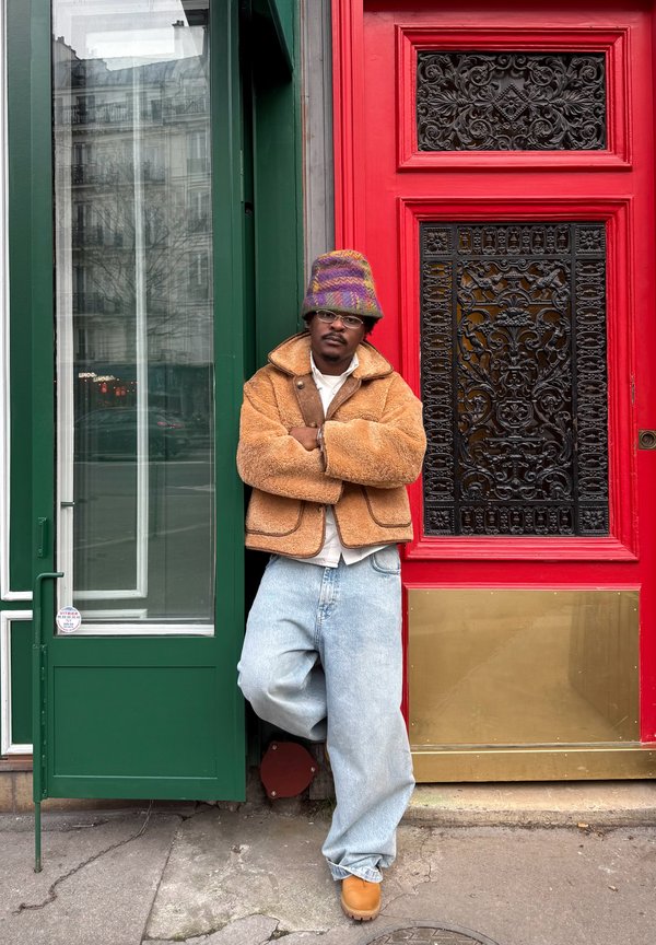 Man wearing tan fleece jacket, light blue jeans, colorful bucket hat, and glasses leaning against a green door beside a red decorative door.