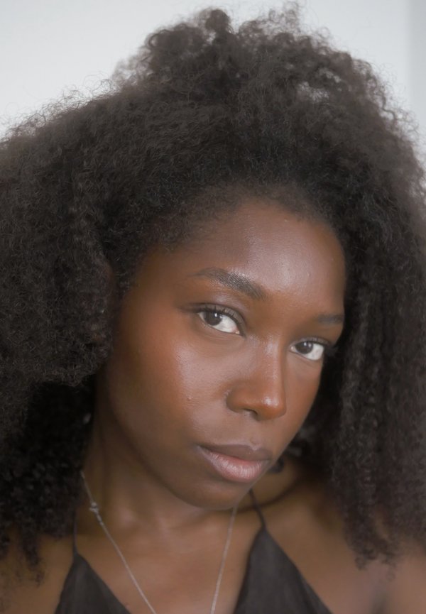 Primo piano di una donna con capelli ricci naturali, pelle scura liscia, che indossa un top nero e una sottile collana d'argento, guardando leggermente di lato.