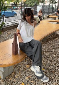 Jeune femme en haut blanc et pantalon gris assise sur un banc en bois courbé à l'extérieur, avec un sac marron à ses côtés sur le sol en gravier.