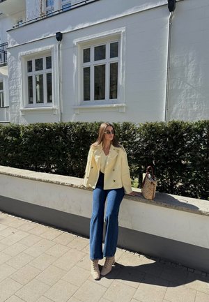 Podwójnie zapinana żółta kurtka, niebieskie dzwony, beżowe sznurowane obcasy, a także pleciona torebka, siedząca w oparciu o kamienny mur.