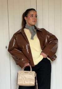 Brown glossy leather jacket, cream knitted jumper, grey scarf, black trousers, holding a small beige handbag with top handles.