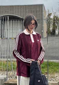 Mørkerød sportsjakke med krage, hvite vertikale striper og blonder, brukt over hvite bukser; svart skulderveske holdt foran.