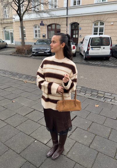 Mujer joven con cabello oscuro recogido en una coleta, vistiendo un suéter marrón y crema a rayas, falda oscura, botas, y sosteniendo un bolso marrón en una calle de la ciudad.