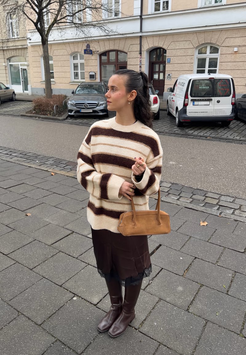 Jeune femme aux cheveux bruns attachés en queue de cheval, portant un pull rayé marron et crème, une jupe foncée, des bottes, tenant un sac à main marron dans une rue de la ville.