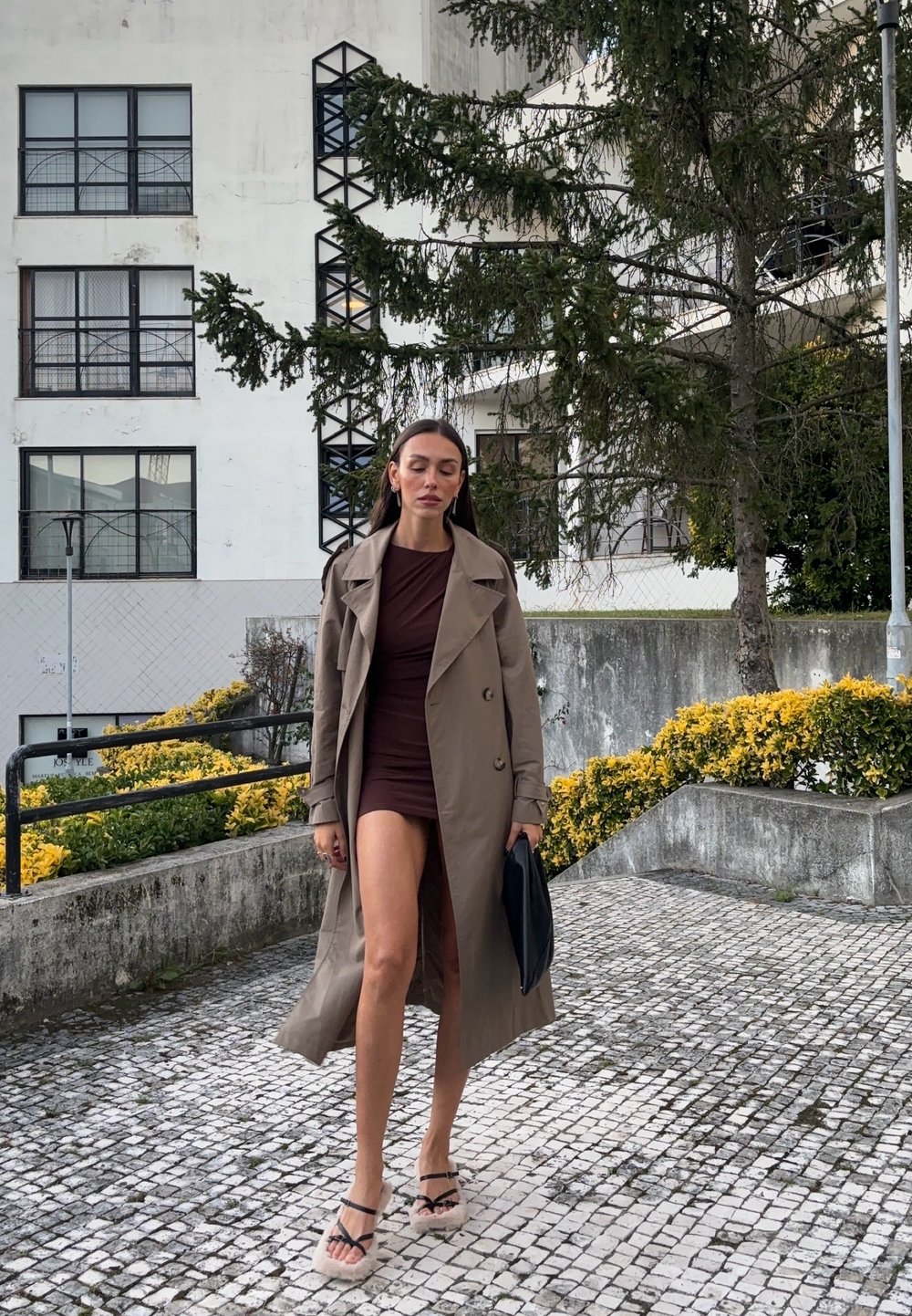 Manteau beige sur une robe brune ajustée, sac à main noir, et sandales à lanières, sur fond de verdure et de pavé carrelé.