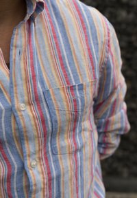 Cămașă cu nasturi, decorată cu dungi verticale în nuanțe de albastru, roșu și galben. Construită dintr-un material ușor, cu un singur buzunar pe piept și mâneci suflecate.