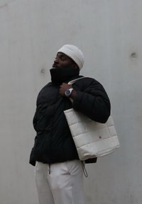 Giacca a vento nera, berretto bianco, che tiene una borsa a tracolla di pelle chiara. Orologio da polso notevole e pantaloni bianchi visibili; texture liscia in tutto.