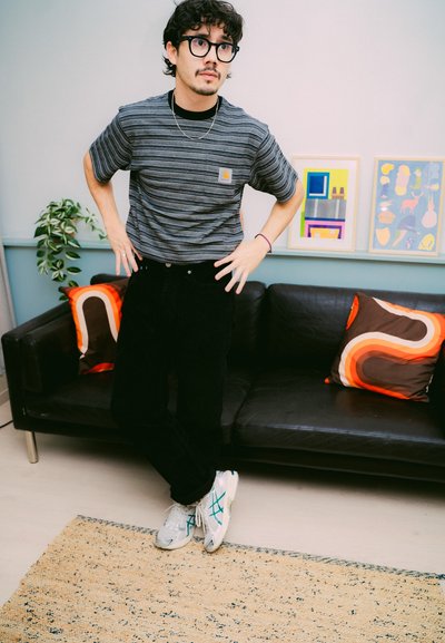 Un hombre con gafas y camisa a rayas está de pie con las manos en las caderas cerca de un sofá negro con almohadas de patrón retro y arte enmarcado.