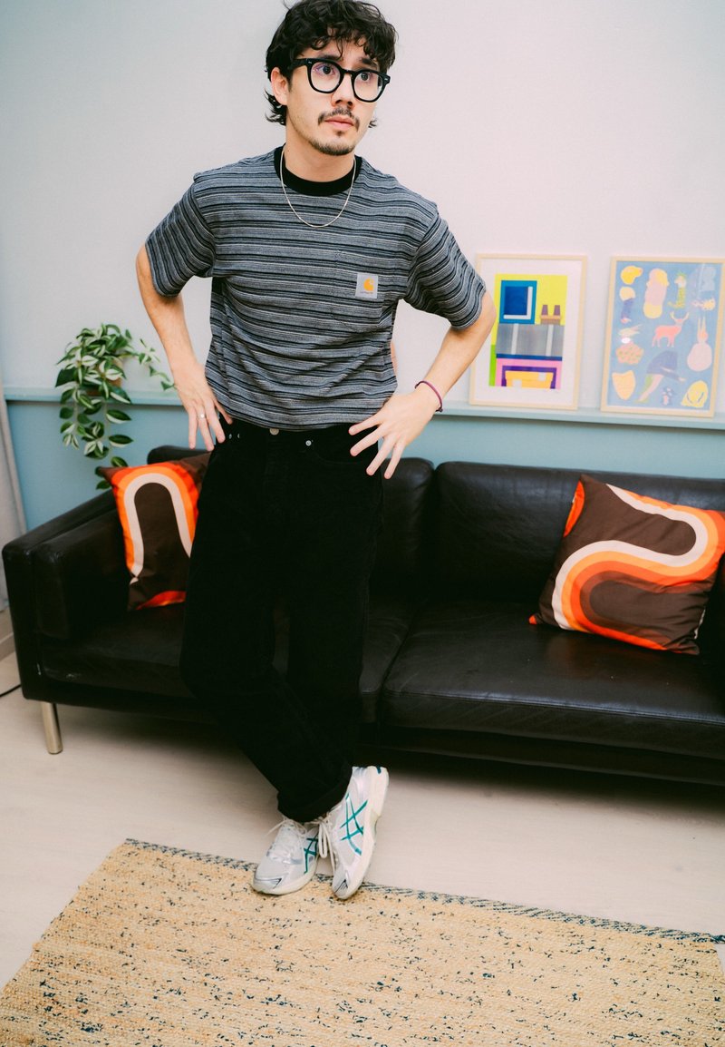 Un homme portant des lunettes et une chemise rayée se tient avec les mains sur les hanches près d'un canapé noir avec des coussins à motifs rétro et des œuvres d'art encadrées.