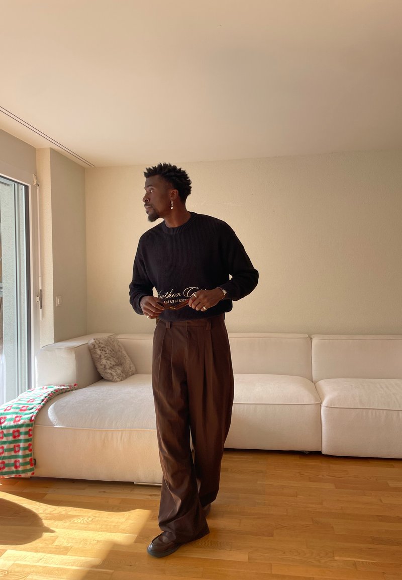 Homme en pull noir et pantalon marron tenant des lunettes, debout près d'un canapé blanc avec une couverture colorée dans un salon baigné de soleil.
