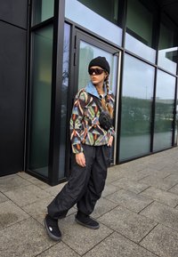 Casaco de fleece com padrão geométrico multicolorido, gola azul; calças oversized pretas; gorro preto; óculos de sol escuros; pequena mala preta.