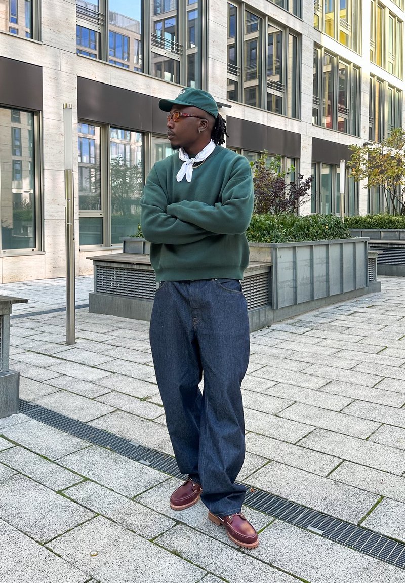 Ανεπίσημη εμφάνιση με σκούρο πράσινο πουλόβερ, ανοιχτό πόδι σκούρων τζιν παντελονιών, μαροονιές λουστρίνια και πράσινο καπέλο με λευκή μαντήλα. Αστική τοποθεσία.