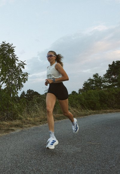 Zapatillas de correr con acentos azules, pantalones cortos negros y una camiseta sin mangas ligera. El corredor lleva gafas de sol y un reloj de fitness, en un camino de grava.