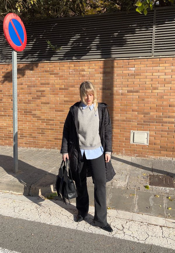 Vrouw die op een stoep staat bij een parkeerverbodsbord, gekleed in een zwarte jas, grijze trui, blauw hemd, zwarte broek en een zwarte handtas vasthoudt.