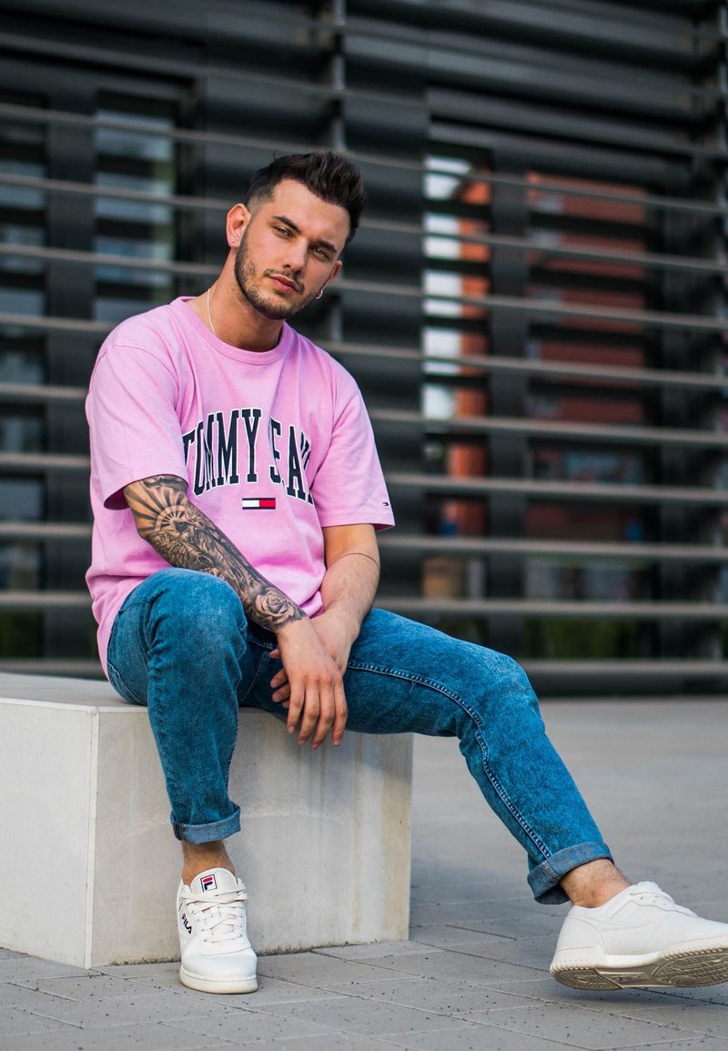 Rosanes Baumwoll-T-Shirt mit "TOMMY"-Logo, blaue Jeans, und weiße Sneakers mit Branding. Armtattoo sichtbar, sitzt auf einer hellen Steinplattform.