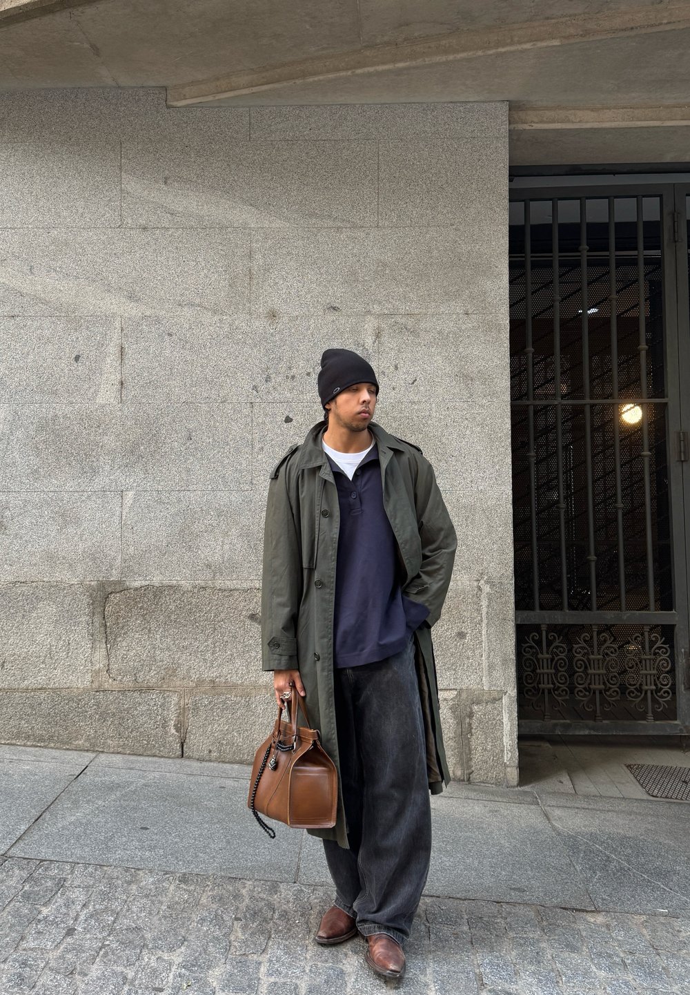 Μακρύ ελαιόχρωμο παλτό, ναυτικό πόλο και φαρδιές μαύρες παντελόνες. Καφέ τσάντα χειρός και καφέ παπούτσια. Γκρι πέτρινος τοίχος και μεταλλική πύλη στο βάθος.