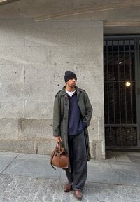 Μακρύ ελαιόχρωμο παλτό, ναυτικό πόλο και φαρδιές μαύρες παντελόνες. Καφέ τσάντα χειρός και καφέ παπούτσια. Γκρι πέτρινος τοίχος και μεταλλική πύλη στο βάθος.