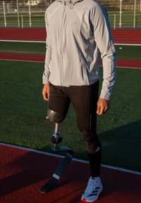 Sportlane, kellel on põlve alt prostetiline jalg, seisab jooksurajal, kandes kerget jopet, musti retuuse ja valgeid spordijalatsid.