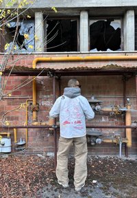 Gray jacket with hood, featuring red graffiti-like patterns on the back. Beige cargo pants, standing by exposed pipes against brick wall.