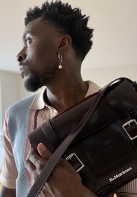 Homme aux cheveux texturés, portant une boucle d'oreille en perle et un tatouage au cou, tient un sac en cuir brun foncé Dr. Martens sur l'épaule à l'intérieur.