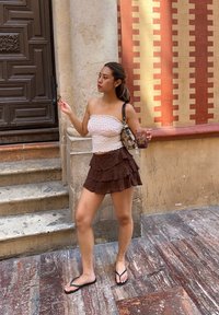 Strapless vit topp med rosa mönster, brun skurna rynkig kjol, svarta flip-flops, håller en dryck, står nära stentrappor.