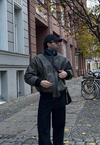 Jacket bomber cinza em pele sintética com fecho, usada sobre um suéter azul de gola alta, combinada com calças pretas e uma bolsa preta, em um ambiente urbano.