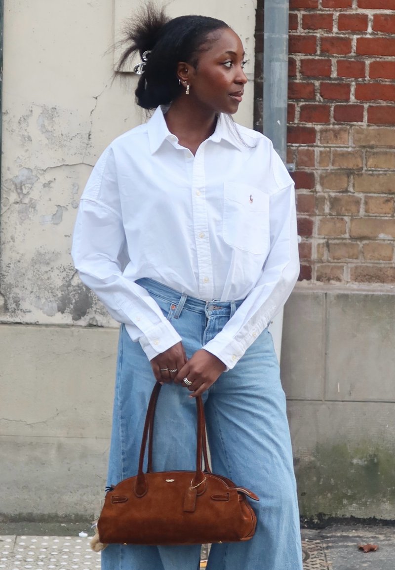 Chemise blanche à col avec une poche, jean à jambes larges bleu clair et sac à main en daim marron, doté de poignées rondes et d'une fermeture éclair.