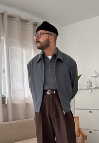 Vyras su barzdą ir akiniais, vilkintis juodą beanie, pilką švarką, tamsiai pilką marškinių, ir rudus kelnes, stovi patalpoje šalia smėlio spalvos užuolaidų.