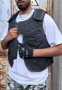 Chaleco negro texturizado con cierre a presión y hombros anchos. También se pueden ver una cadena plateada y guantes negros con nudillos protectores.