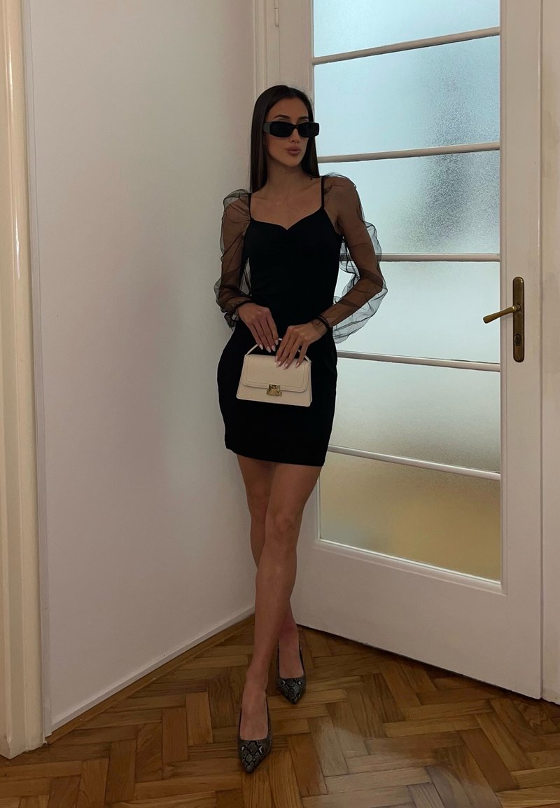Woman in black mini dress with sheer puff sleeves, black sunglasses, patterned heels, holding a small beige handbag, standing by frosted glass door.