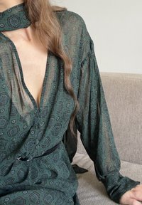 Zielona, przezroczysta bluzka z długimi rękawami, głębokim dekoltem w kształcie litery V i kołnierzykiem typu choker. Posiada wzór paisley w ciemniejszych odcieniach oraz zapięcie na guziki z przodu.