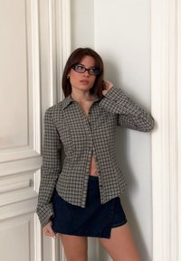 Femme aux cheveux bruns portant des lunettes à monture noire, une chemise à manches longues à carreaux marron, et une jupe bleu foncé, debout à l'intérieur contre des murs blancs.