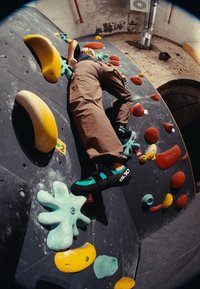Parete da arrampicata con prese di diverse forme color giallo, rosso e turchese; un climber in pantaloni marroni e scarpe da arrampicata nere-verdi sulla parete.
