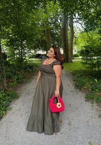 Ελαιόχρωση, χωρίς μανίκια μακριά φόρεμα με βολάν και ραμμένα λεπτομέρειες στο μπούστο, συνδυασμένο με μια φωτειν ή ροζ τσάντα χειρός με λουλούδι διακοσμητικό.