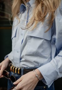 Lyseblå skjorte med knapper og to påsydde lommer, kombinert med høyhåndte denimjeans og et belte med gulldetaljer.