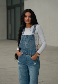 Peto de mezclilla en un lavado claro con acentos desgastados, combinado con una camiseta ajustada de manga larga blanca, con las manos en los bolsillos, presentando un diseño elegante.