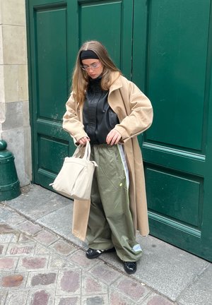 Mujer joven con abrigo largo beige, chaqueta negra, pantalones anchos verde oliva, zapatos negros y gafas, sosteniendo un bolso beige junto a una puerta verde.