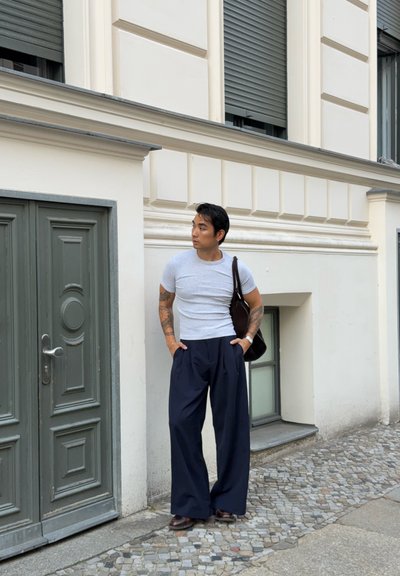 Camiseta gris ajustada, pantalones anchos de rayas marino, zapatos marrones y un bolso marrón de hombro. Delante de una puerta verde y un edificio de color claro.