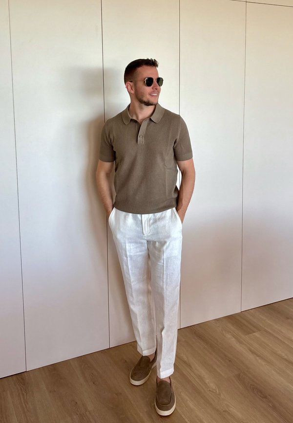 Homme portant des lunettes de soleil, un polo en maille marron à manches courtes, un pantalon en lin blanc et des chaussures marron à enfiler, debout avec les mains dans les poches.
