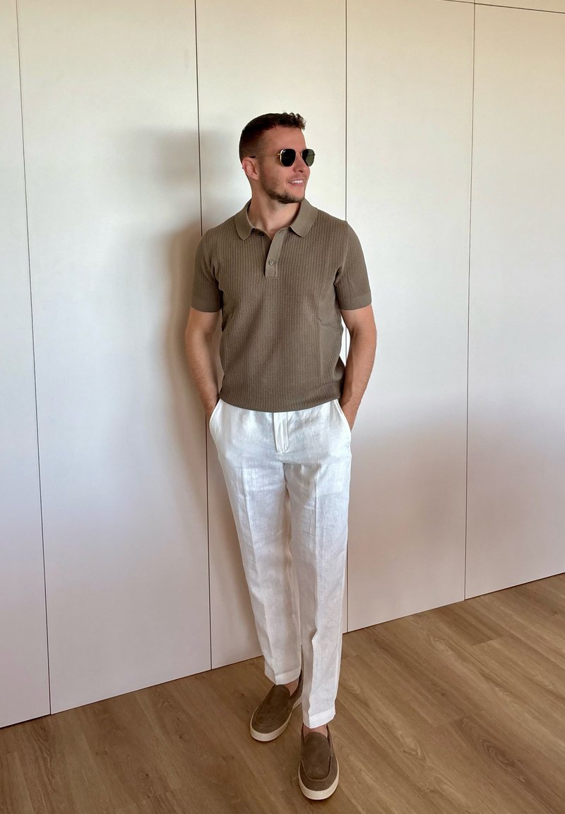Homme portant des lunettes de soleil, un polo en maille marron à manches courtes, un pantalon en lin blanc et des chaussures marron à enfiler, debout les mains dans les poches.