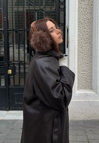 Femme portant des cache-oreilles en fourrure marron, une veste en cuir noire et un pull blanc, se tenant près d'une grille en fer noire et d'un mur blanc texturé.