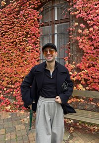 Person som har på seg en svart frakk, svart topp og grå plisserte bukser. Tilbehøret inkluderer en svart hatt og solbriller. Veggen i bakgrunnen har røde blader.