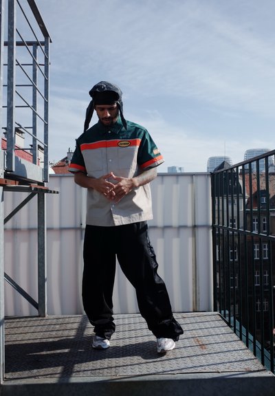 Camisa de manga corta con paneles verdes y beige, detalles en naranja, dos bolsillos en el pecho, combinada con pantalones negros holgados y zapatillas blancas.