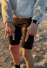 Pantaloncini sportivi neri con un design elegante caratterizzato da un motivo ondulato. Abbinati a una giacca leggera, riflettente, con cerniera. Mani che compiono un gesto.